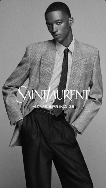 Black male fashion model poses in a light gray tweed suit, dark shirt, and tie for Saint Laurent Men's Spring 25 advertisement.