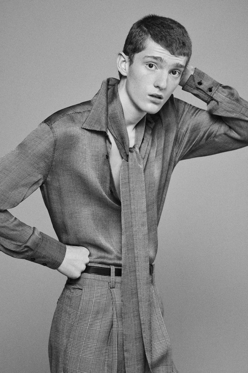 Studio portrait of a young adult male wearing a gray shirt with a tie and patterned pants, posing against a gray backdrop.