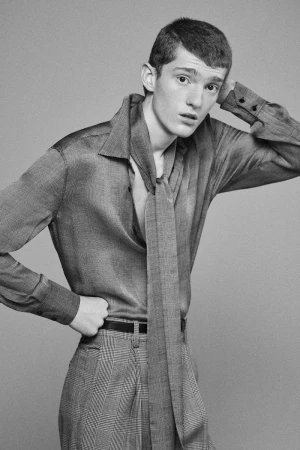 Studio portrait of a young adult male wearing a gray shirt with a tie and patterned pants, posing against a gray backdrop.