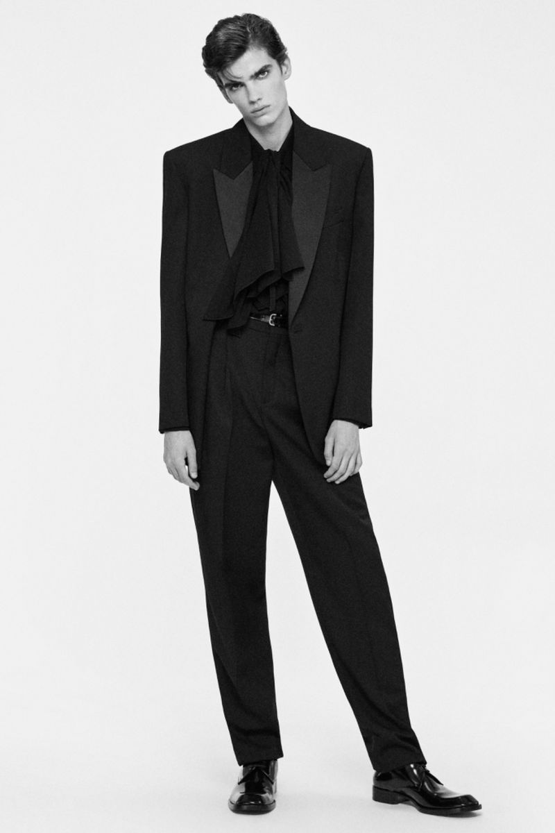 Black and white fashion portrait of a male model wearing a black tuxedo and tie posing in a photo studio against a plain backdrop.