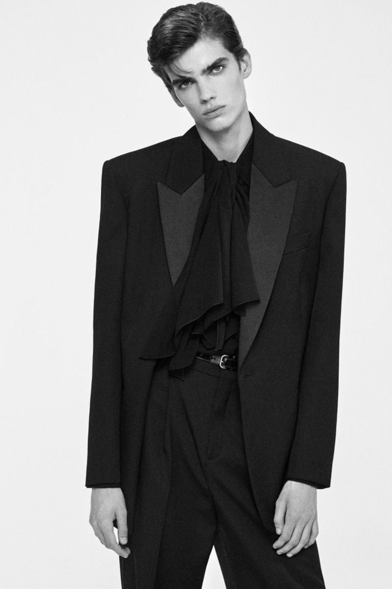 Black-and-white fashion portrait of a slim, fair-skinned male model wearing a dark ruffled shirt, trousers, and blazer against a white backdrop.