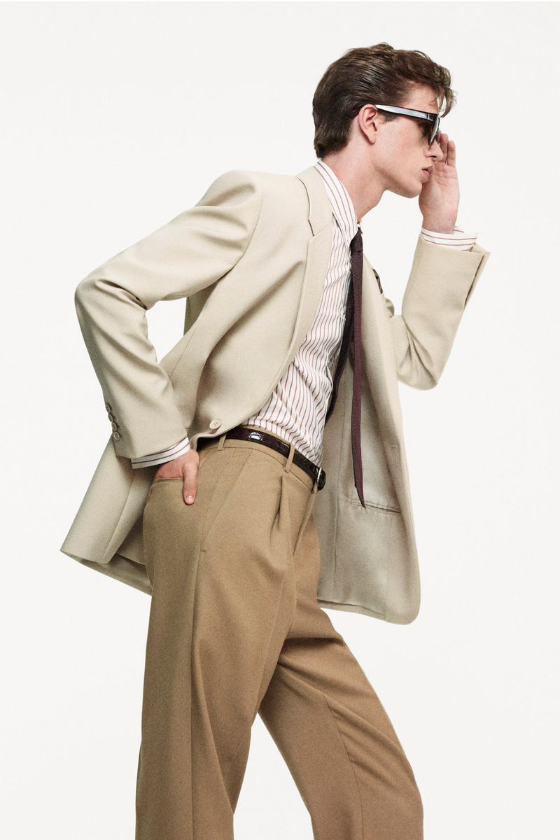Side-view shot of a man wearing sunglasses, a light-colored blazer, a striped dress shirt, a dark tie, and brown trousers in a studio.