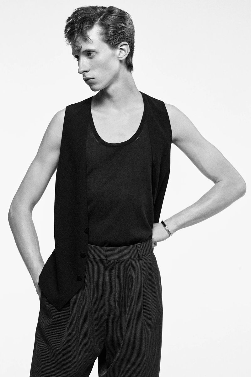 Monochrome portrait of a slender man in a black tank top and vest, with pinstripe pants, standing against a stark white backdrop.