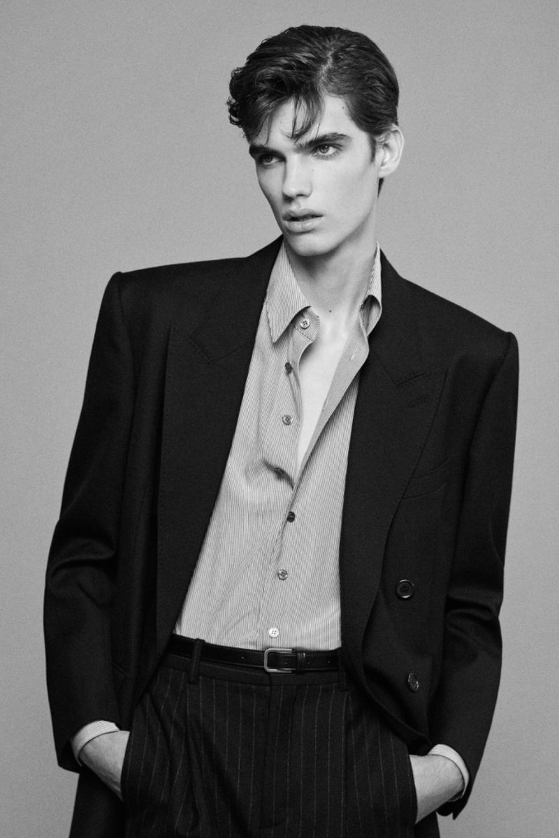 Monochrome portrait of a young male model wearing a tailored dark blazer and buttoned shirt against a grey studio backdrop.