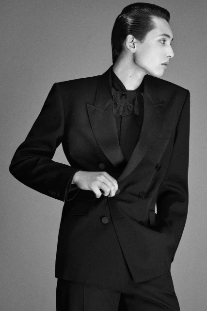 Monochrome studio portrait of a person in a dark double-breasted suit looking left with slicked-back hair against a neutral background.