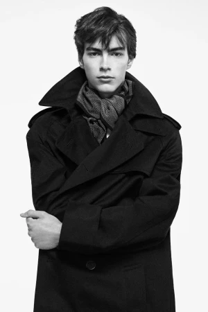 Monochrome portrait of a male model wearing a dark trench coat and patterned scarf against a plain white background.