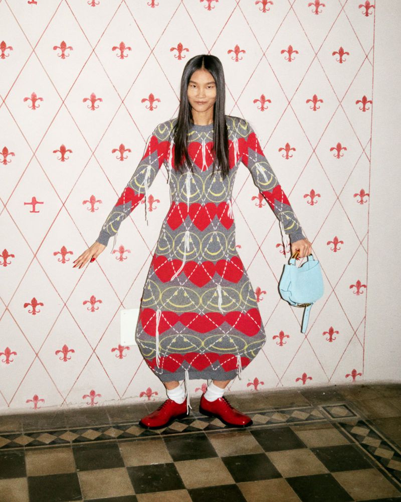 Asian female in a patterned gray, red, and yellow sweater dress, with a light blue purse and red shoes, standing in front of wallpaper.