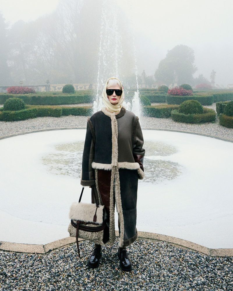 Fashion shot of an adult female in a coat and headscarf holding a purse standing in front of a fountain with hedges in the background.