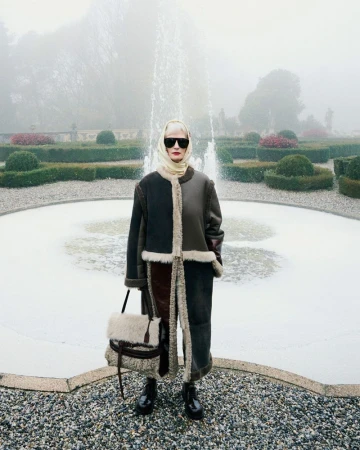 Fashion shot of an adult female in a coat and headscarf holding a purse standing in front of a fountain with hedges in the background.