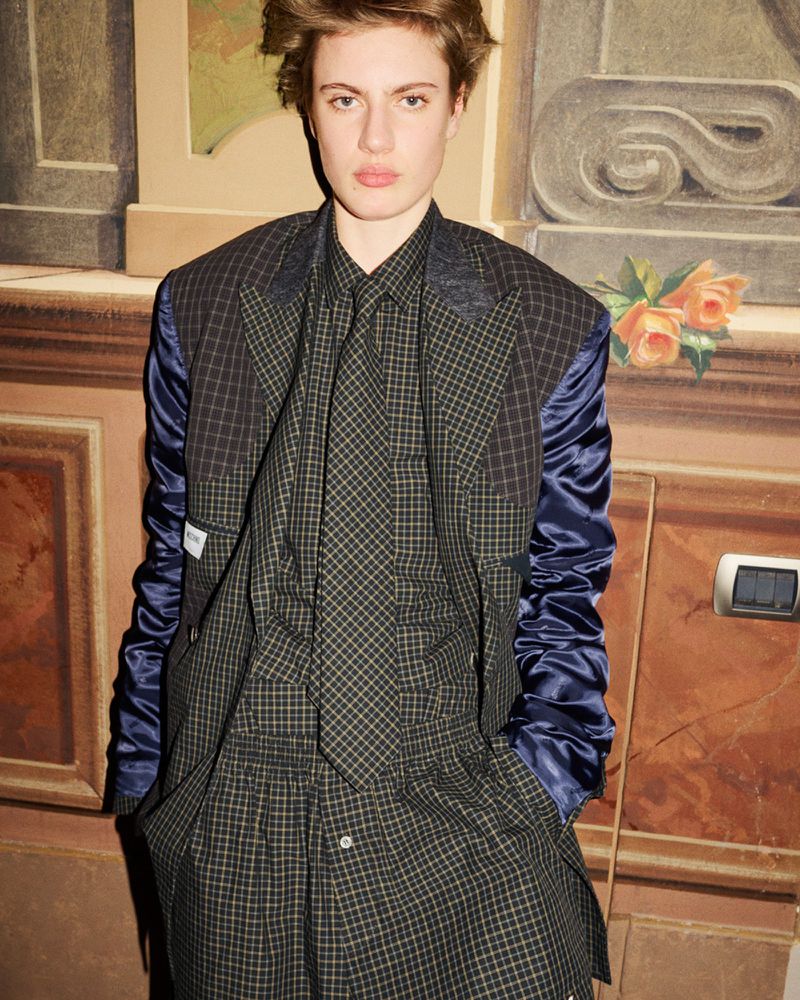 An adult female poses indoors wearing a plaid blazer with blue sleeves, a matching button-down shirt, and tie with a neutral expression.
