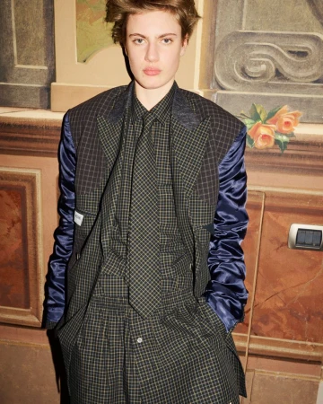 An adult female poses indoors wearing a plaid blazer with blue sleeves, a matching button-down shirt, and tie with a neutral expression.