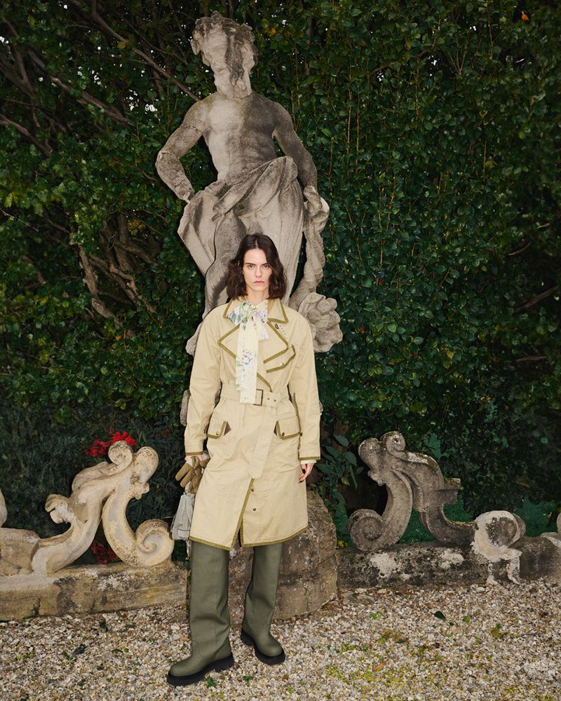 A woman in a beige trench coat and green boots stands in a garden with a stone statue and lush green foliage in the background.