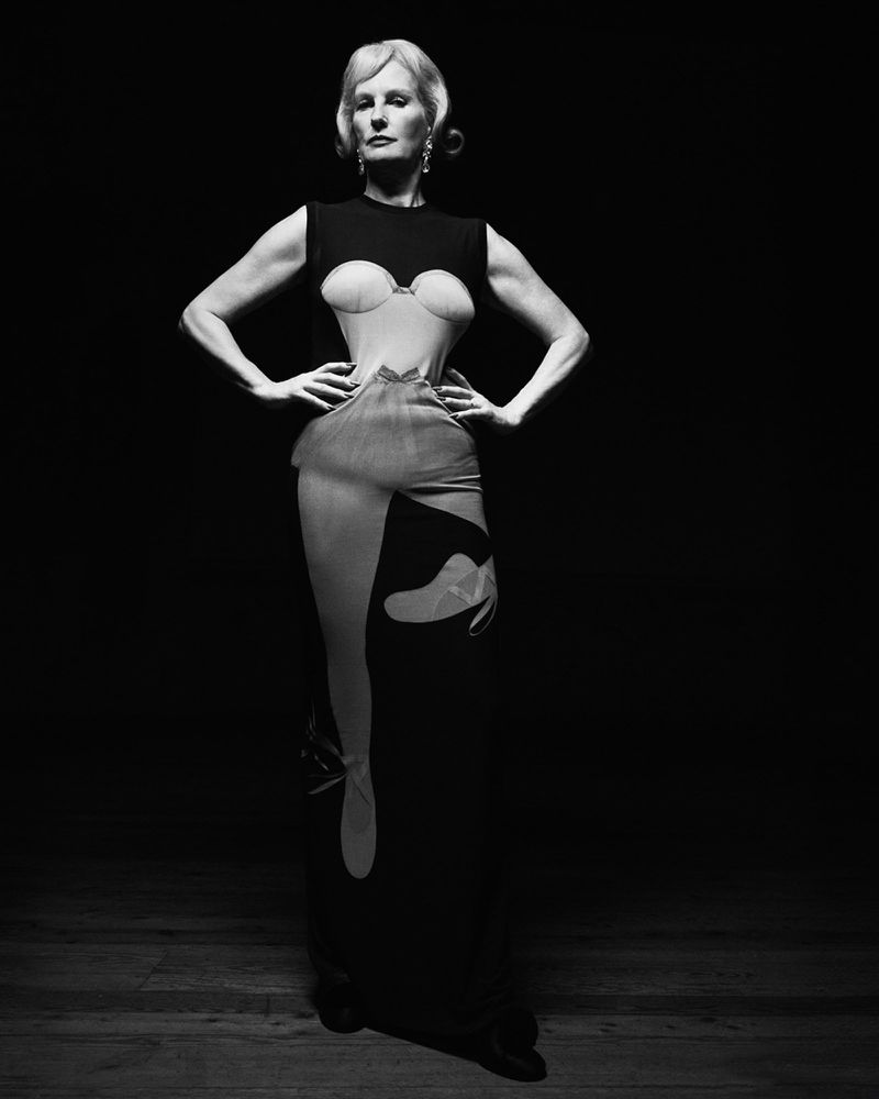 Elegant woman with blonde hair poses in a unique black dress, striking a powerful stance in a studio setting with dark backdrop.