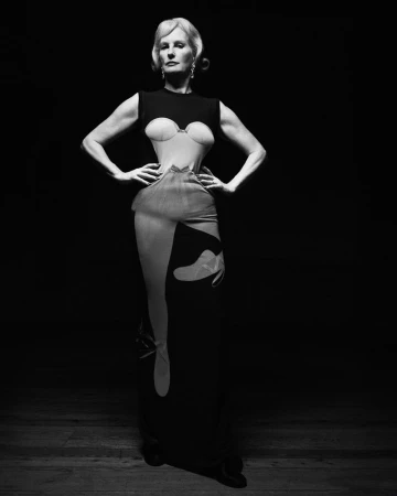 Elegant woman with blonde hair poses in a unique black dress, striking a powerful stance in a studio setting with dark backdrop.