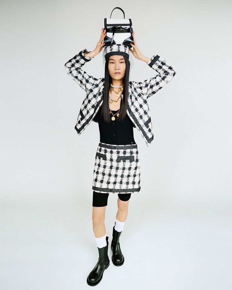 A full shot of a person wearing a black and white plaid outfit and black boots, holding a small bag on their head against a white background.
