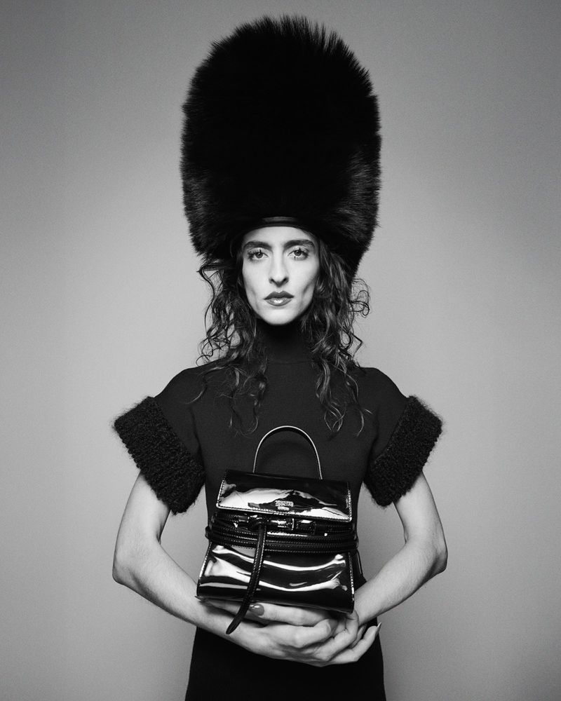 Woman in black dress and large fur hat holds black patent leather handbag in studio portrait against grey background. Fashion editorial.