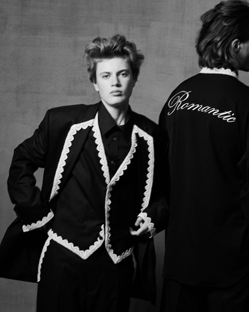 Two people in black outfits with white trim and the word 'Romantic' embroidered on the back, stand against a neutral wall.