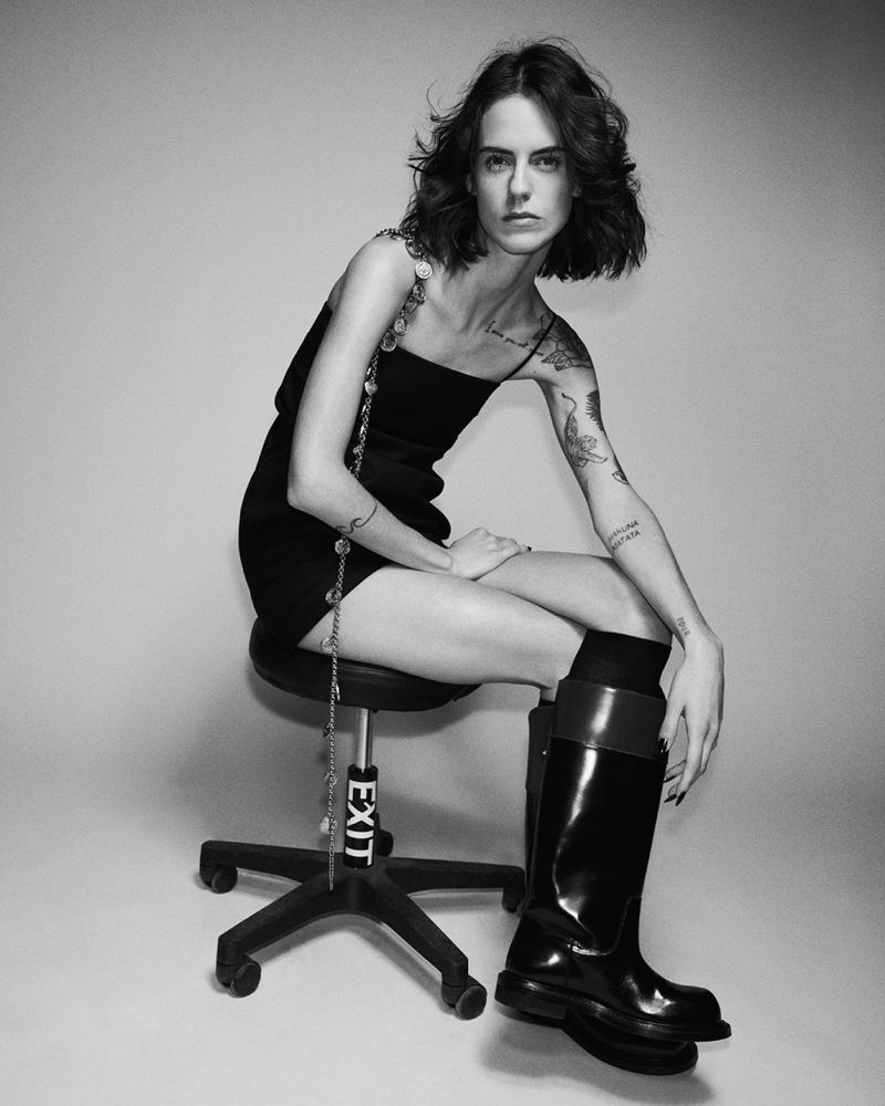 Monochrome portrait of a fair-skinned woman with tattoos, seated on a chair, wearing a black dress and black shiny boots, indoor studio.