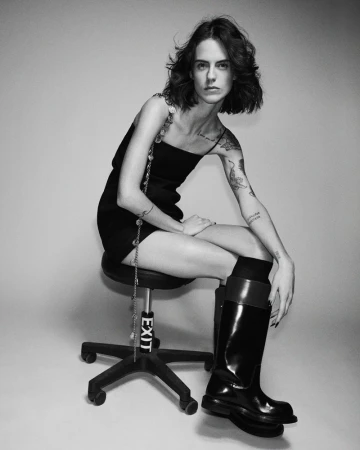 Monochrome portrait of a fair-skinned woman with tattoos, seated on a chair, wearing a black dress and black shiny boots, indoor studio.
