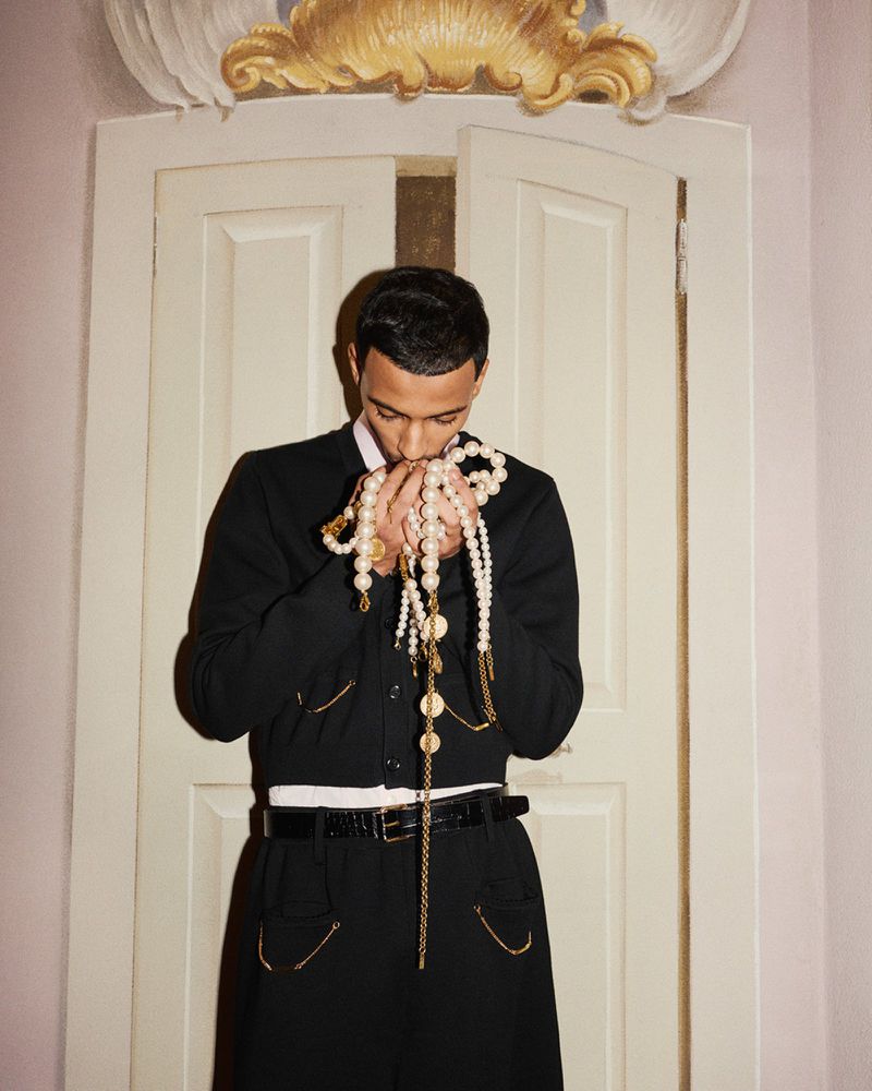 Portrait of a Black man indoors holding strands of pearl and gold beaded necklaces, wearing a black jacket and skirt suit.