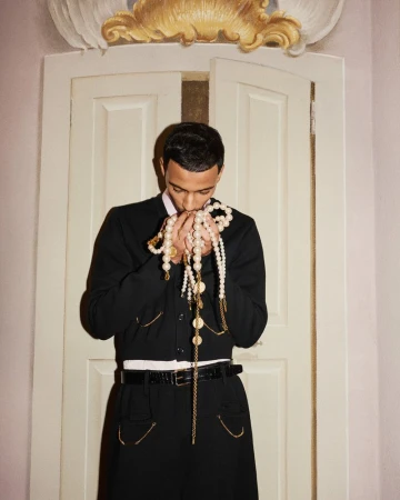 Portrait of a Black man indoors holding strands of pearl and gold beaded necklaces, wearing a black jacket and skirt suit.