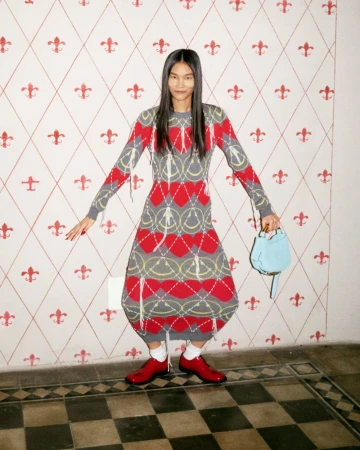 Model wearing a patterned knit dress with hearts and smiley faces holding a blue handbag against a tiled wall
