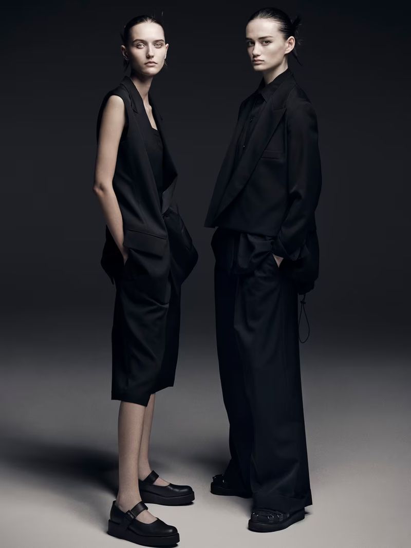 Two women in black oversized fashion, one in vest and culottes, the other in jacket and trousers, stand in a neutral studio.