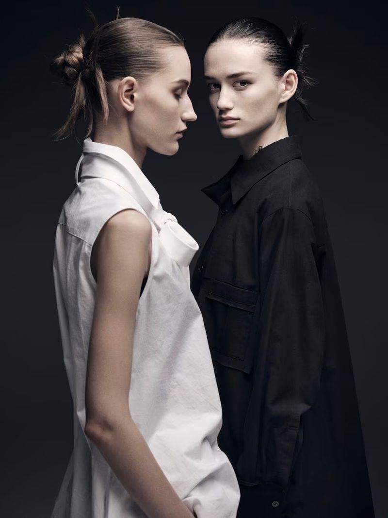 Two women in contrasting black and white shirts pose in a studio with soft lighting, highlighting their similar features and minimalist fashion.