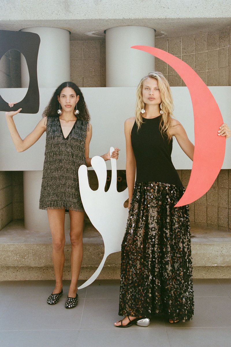 Two women pose in dresses, holding large geometric shapes in front of a concrete wall with cylindrical columns, fashion photography.
