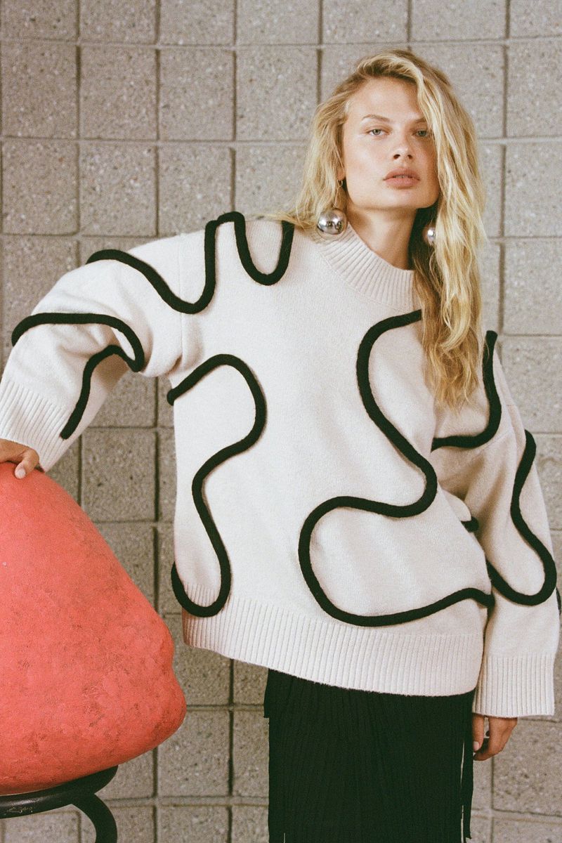 A woman in a cream sweater with a black wavy pattern poses in front of a textured gray block wall with a coral colored sculpture.