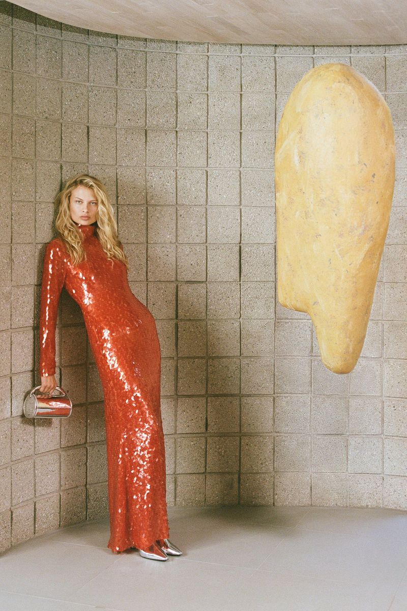 A slender female model in a red sequin gown stands against a textured block wall in a studio, holding a silver bag, for a fashion editorial.