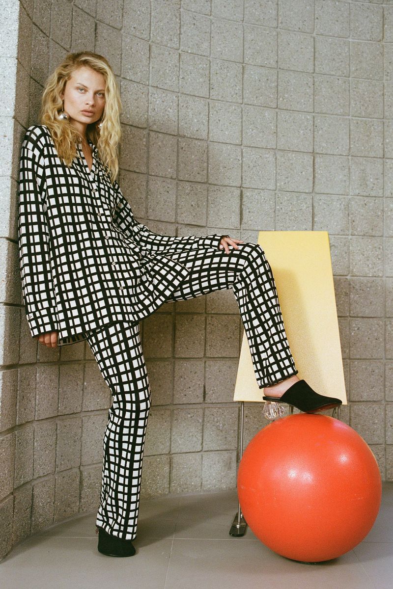 A blonde-haired adult female is posing in a checkered print lounge set and black mules in an indoor studio against a tiled wall.