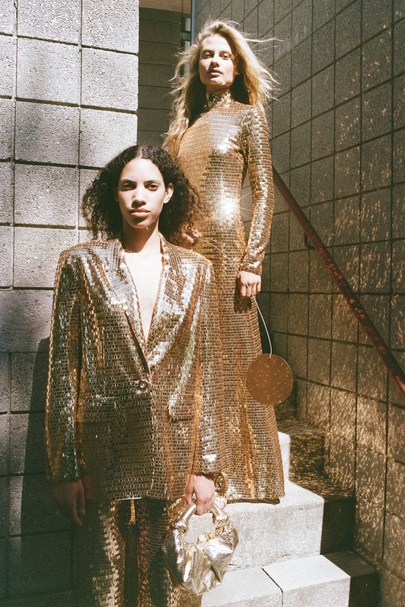 Two fashion models in gold sequin outfits pose on stairs. One wears a blazer and pants, and the other wears a long-sleeved gown.