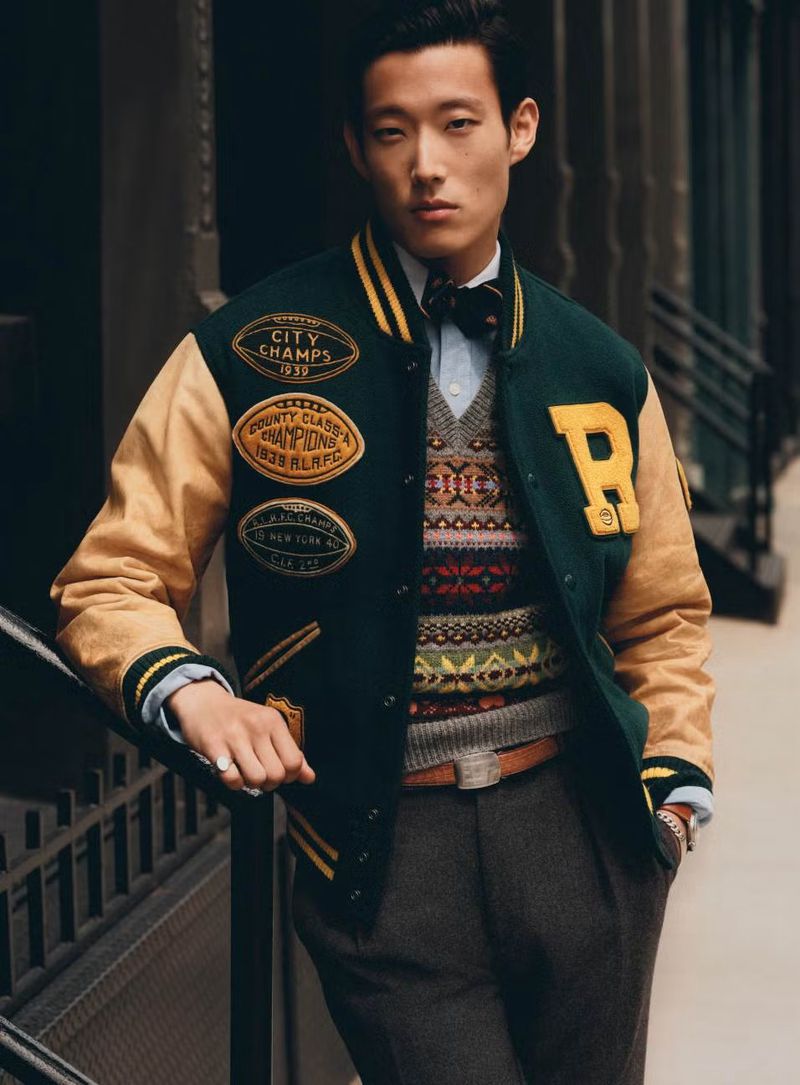 Man in letterman jacket and patterned sweater posing in an urban setting with brick walls and metal railings. Fashion advertising.