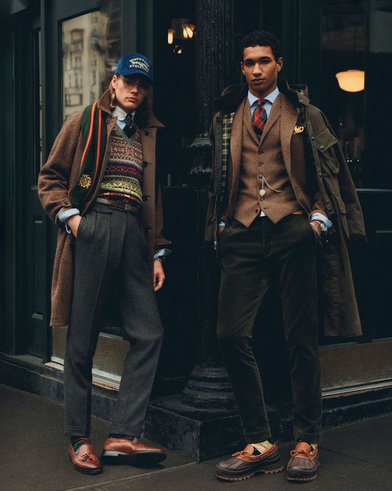 Two men in classic fall fashion stand outdoors in coats, sweaters, dress pants, and dress shoes on a city street, posing for the camera.