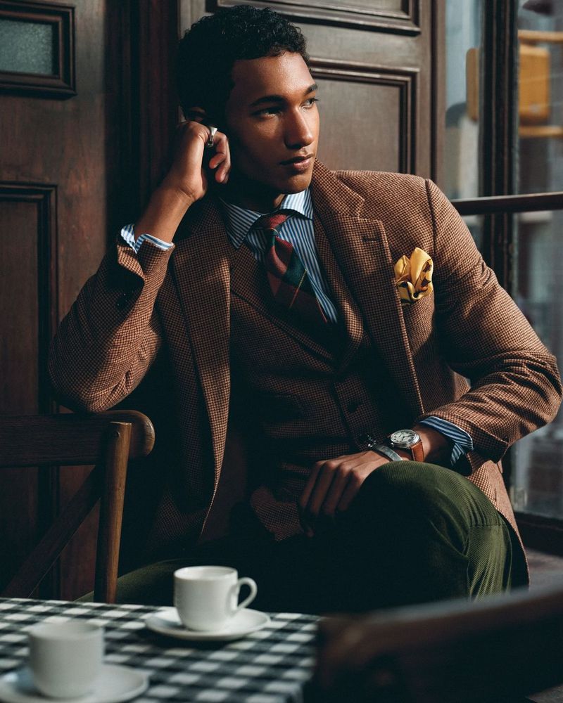 A stylish man in a tweed suit sits by a window with coffee cups, dressed in a vest, tie, and watch for a polished look.