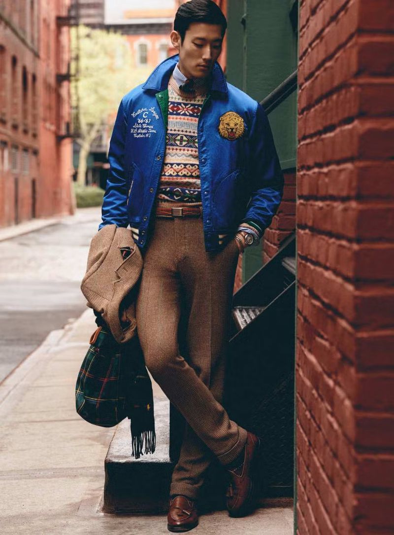 Man in blue jacket with lion emblem posing on a street with brick buildings. He carries a coat and scarf, wearing brown trousers and loafers.