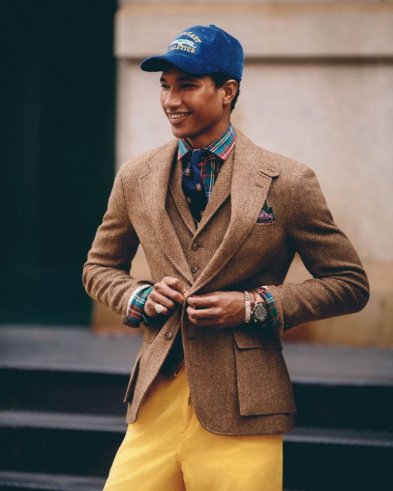Male fashion model smiles, wearing a brown tweed suit, a patterned shirt, and yellow slacks, standing outdoors with a cap.