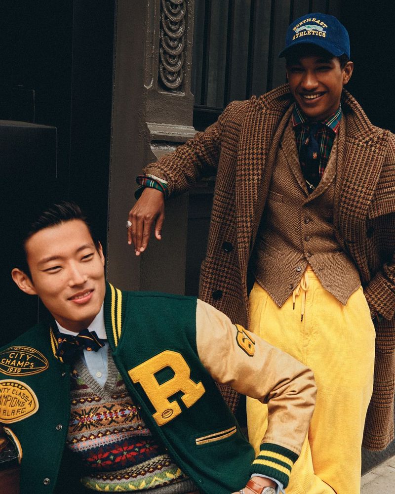 Two men in vintage-style outfits pose outdoors; one wears a letterman jacket and the other is in a tweed coat and baseball cap.