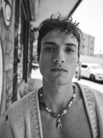 Portrait of a male model wearing a knitted cardigan and beaded necklace against an urban street background in black and white