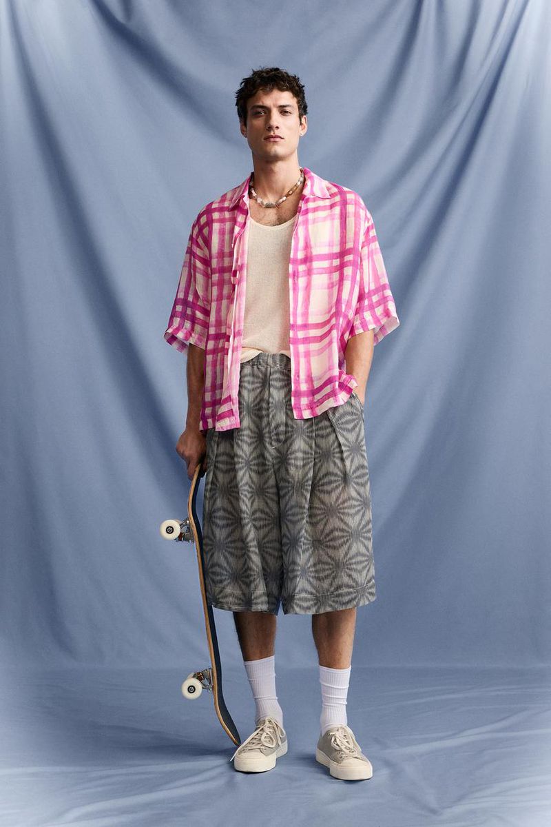 A stylish adult male poses in a pink plaid shirt and patterned shorts, holding a skateboard in a studio setting with a blue backdrop.