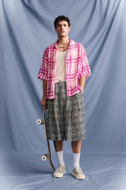 A stylish adult male poses in a pink plaid shirt and patterned shorts, holding a skateboard in a studio setting with a blue backdrop.