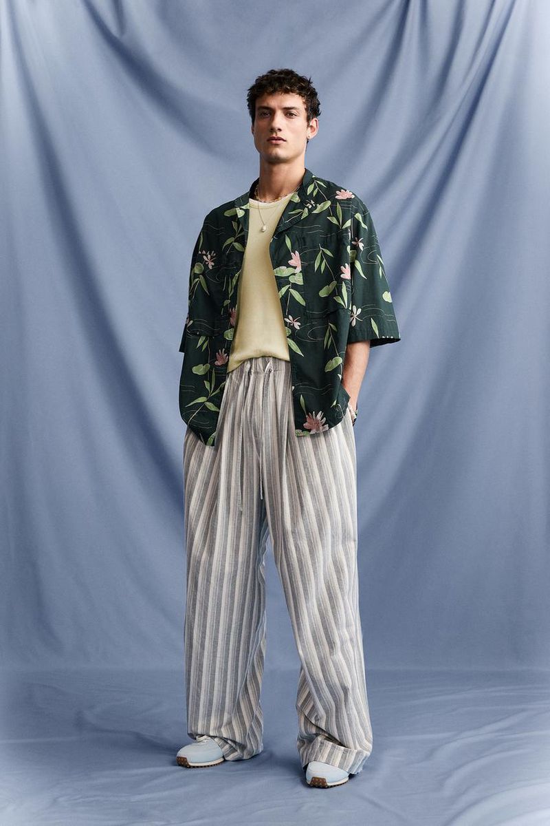 Full-length shot of a male model wearing a floral shirt and striped pants against a blue draped background, posing indoors.