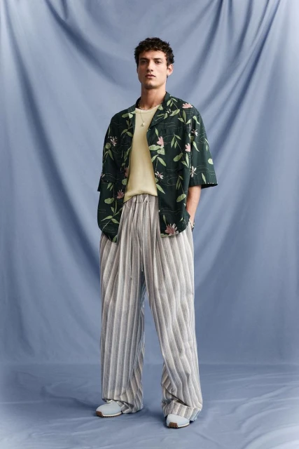 Full-length shot of a male model wearing a floral shirt and striped pants against a blue draped background, posing indoors.