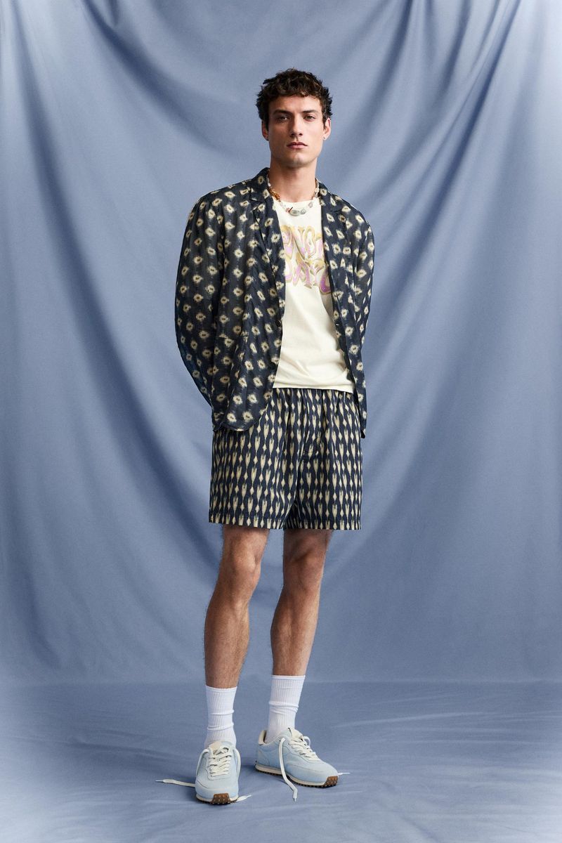 Full-length shot of a young adult male model in a patterned shirt, t-shirt, and shorts, wearing white socks and sneakers, against a blue backdrop.