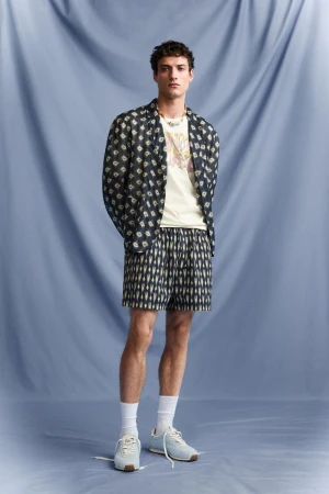 Full-length shot of a young adult male model in a patterned shirt, t-shirt, and shorts, wearing white socks and sneakers, against a blue backdrop.