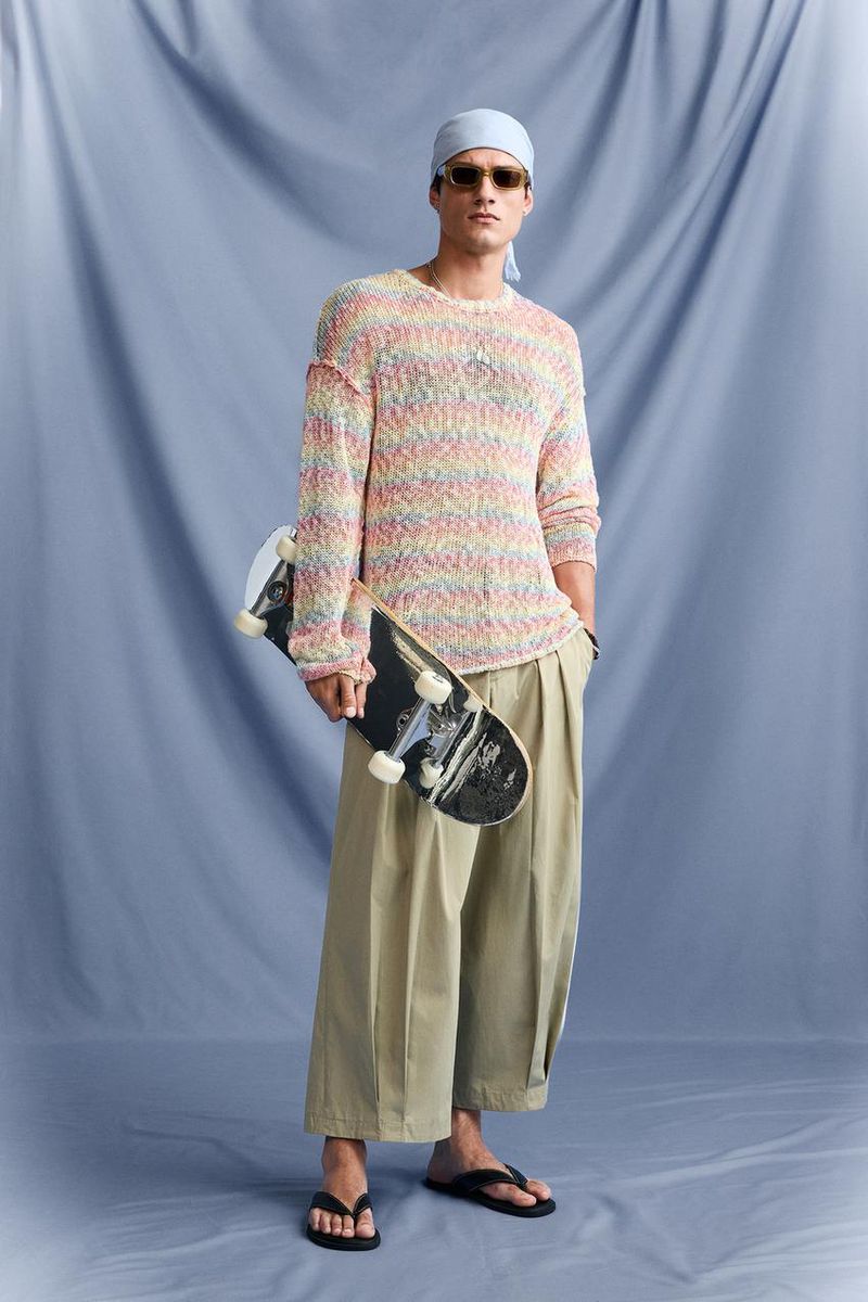 Man in colorful sweater, sunglasses, headscarf, and culottes holds skateboard against a blue studio backdrop. Relaxed fashion editorial.