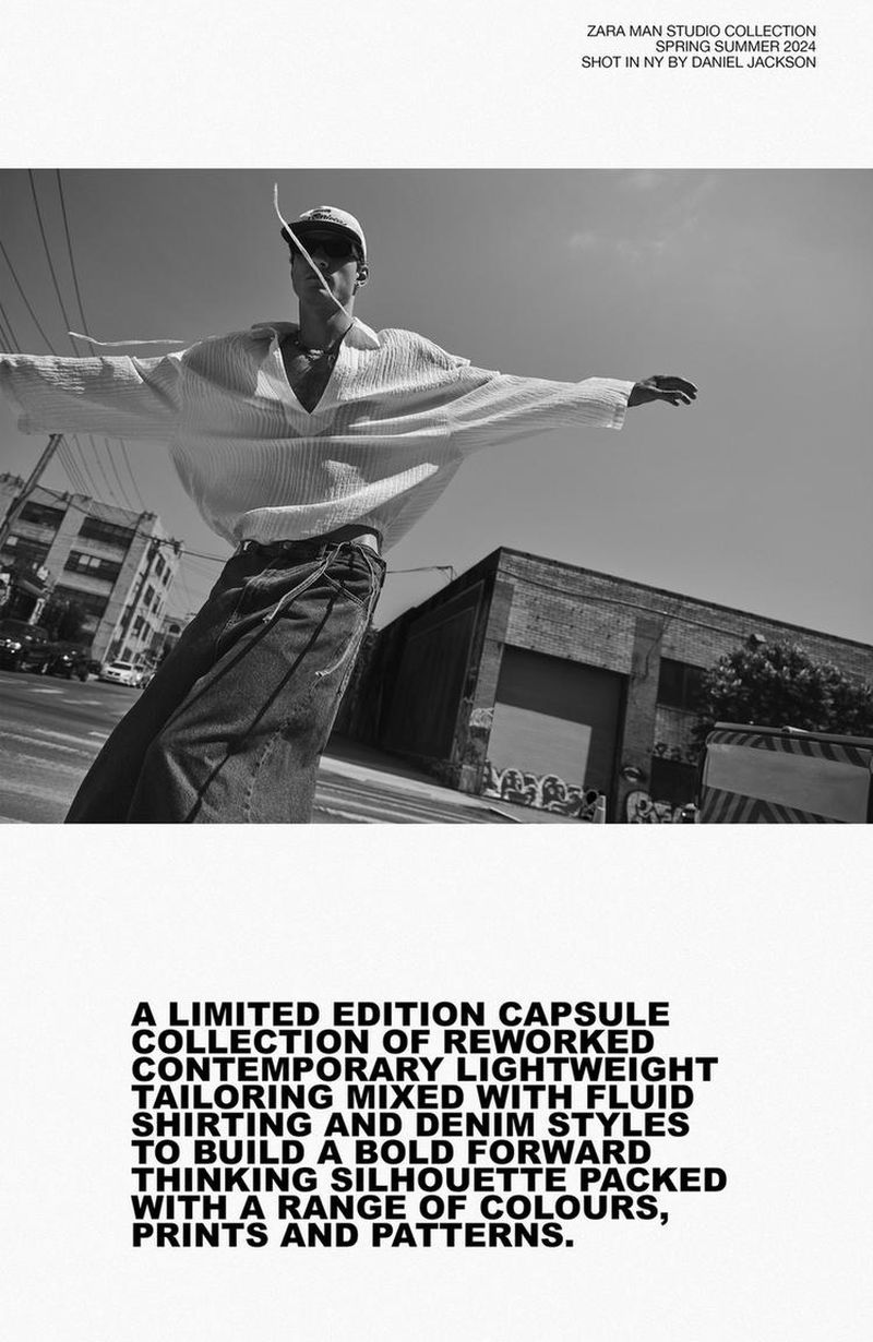 Black and white fashion shot of a man in baggy jeans and a white shirt posing in front of a brick building and sky on a sunny day.