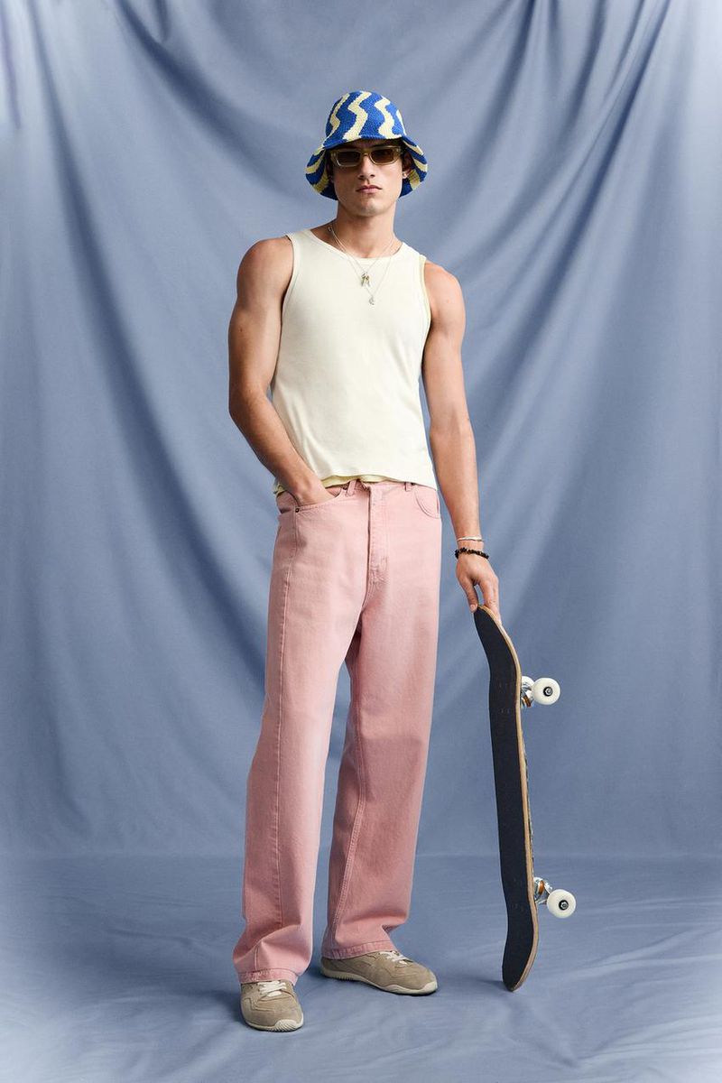 Full shot of a young adult male in a tank top and light pink jeans holding a skateboard in front of a blue backdrop.