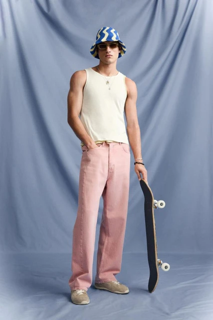 Full shot of a young adult male in a tank top and light pink jeans holding a skateboard in front of a blue backdrop.